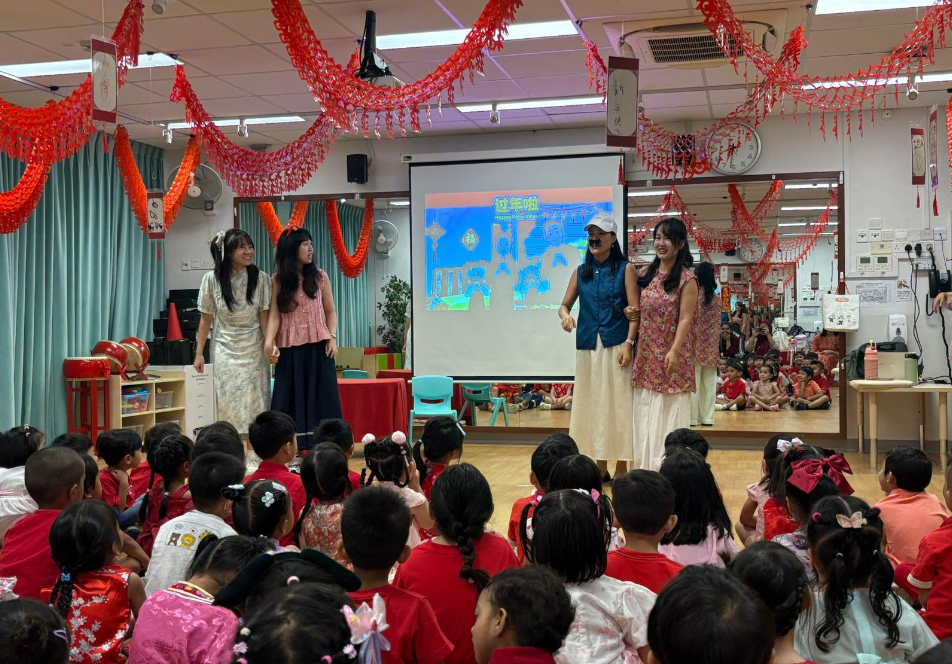 Four women perform on stage before seated children; red streamers hang overhead and a projector screen behind.